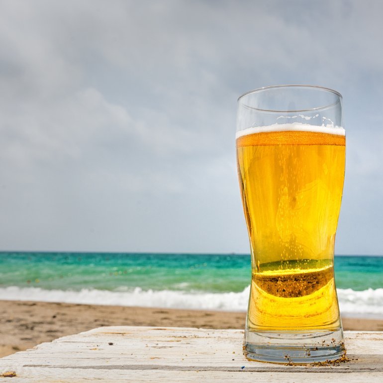 A refreshing glass of beer on a beach, showcasing the vibrant color of ELUSIVE NECTAR surrounded by serene ocean views.
