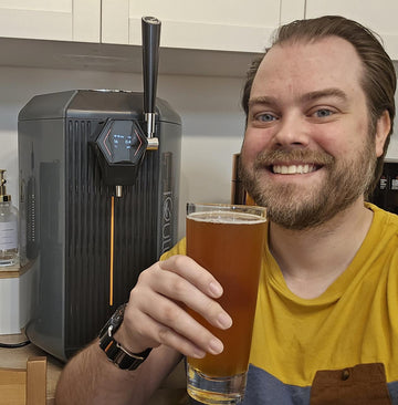 A man smiles while holding a beer in front of a stylish kegerator, showcasing 100 iGulu User Stories.